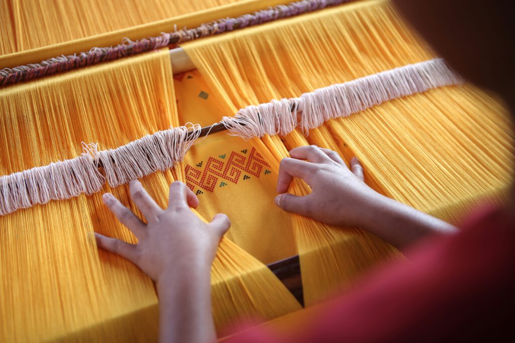 a woman seen from the back who is weaving a yellow piece of cloths by hand on a wooden machine. picture in blog post about the trecena, 13-day forecast, of the mayan calendar for batz.